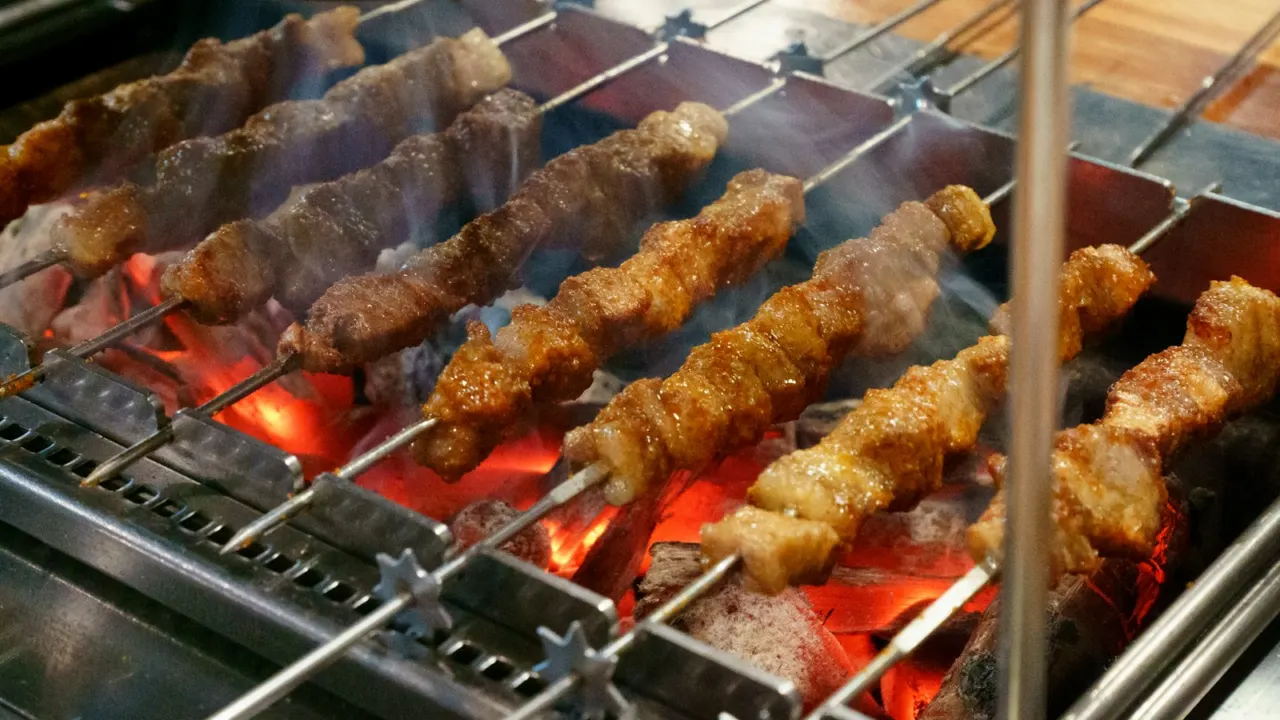 Several skewers of meat cooking side by side on a barbecue grill