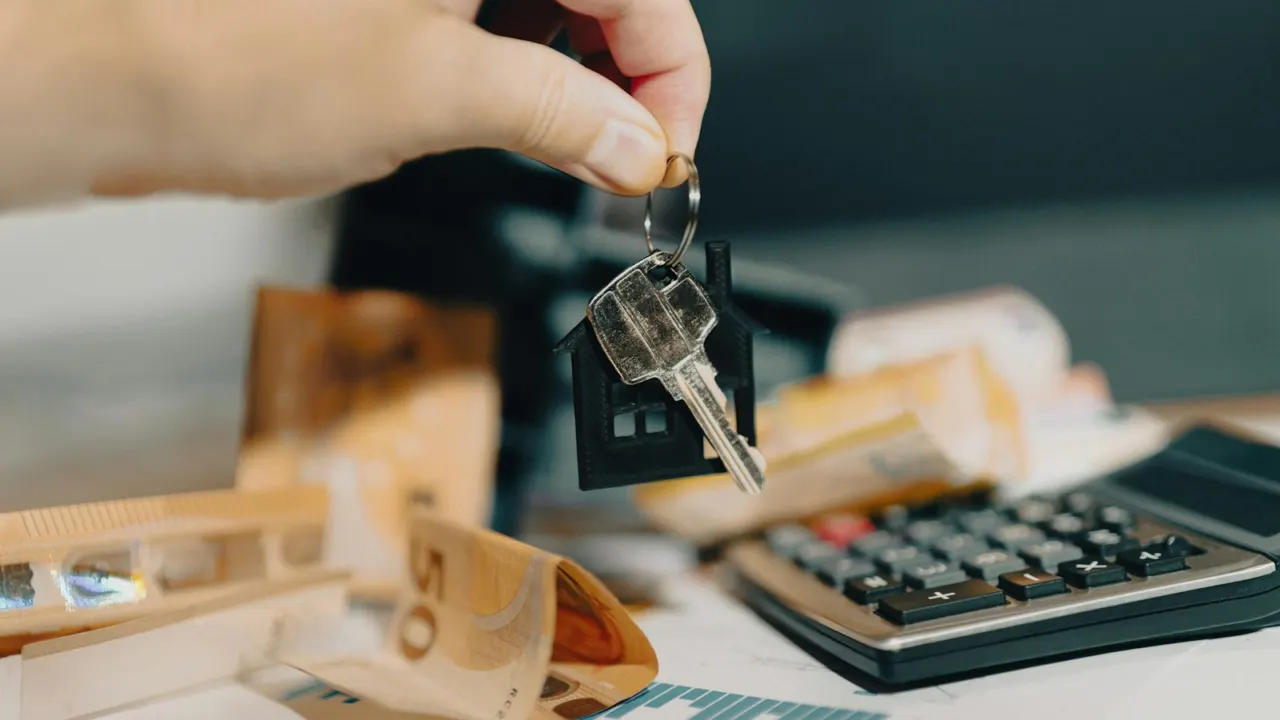 Hand holding a house key on a keychain above a desk with a calculator and paperwork