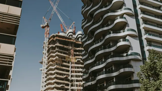 A residential tower under construction with cranes on top, beside a completed tower with curved balconies