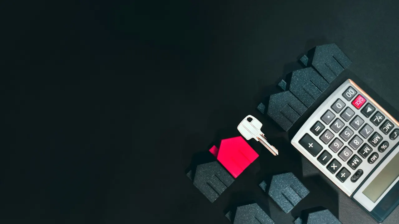 A silver house key, a small red house model and a calculator laid out on a dark desk