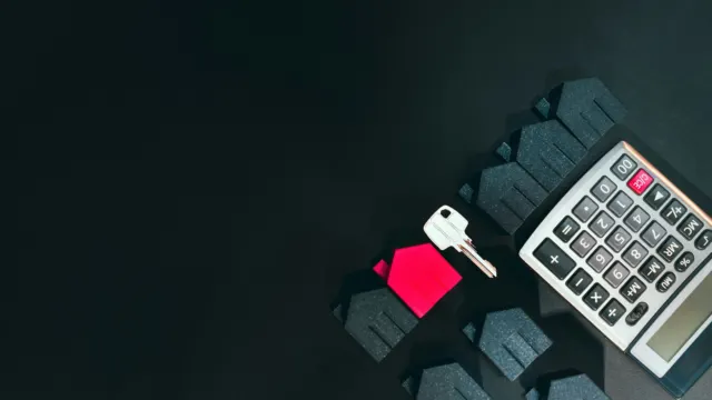 A silver house key, a small red house model and a calculator laid out on a dark desk
