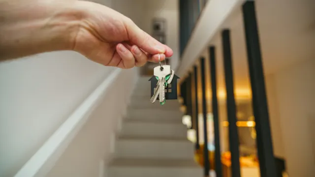 Hand holding a set of keys on a house-shaped keychain in front of a staircase