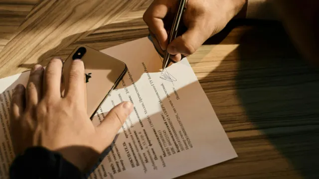 Person signing a printed document with a pen at a wooden desk