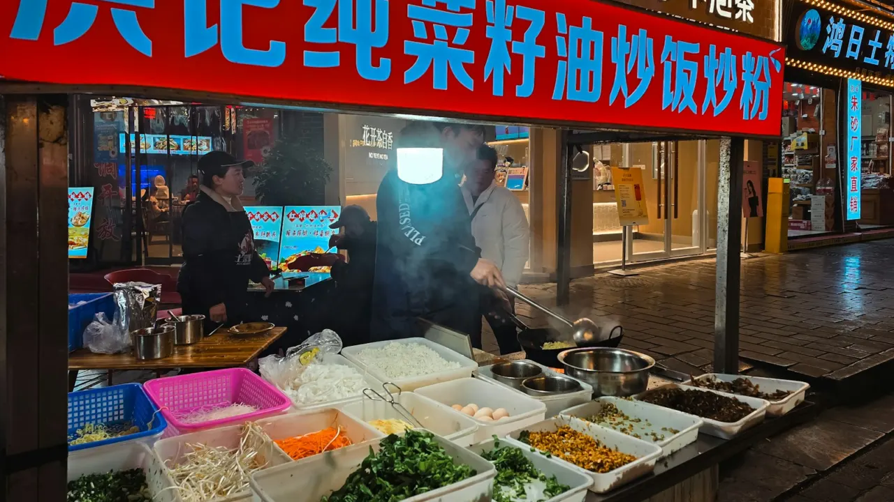 A night market food stall under a red sign, with a cook tossing noodles in a wok
