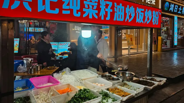 A night market food stall under a red sign, with a cook tossing noodles in a wok