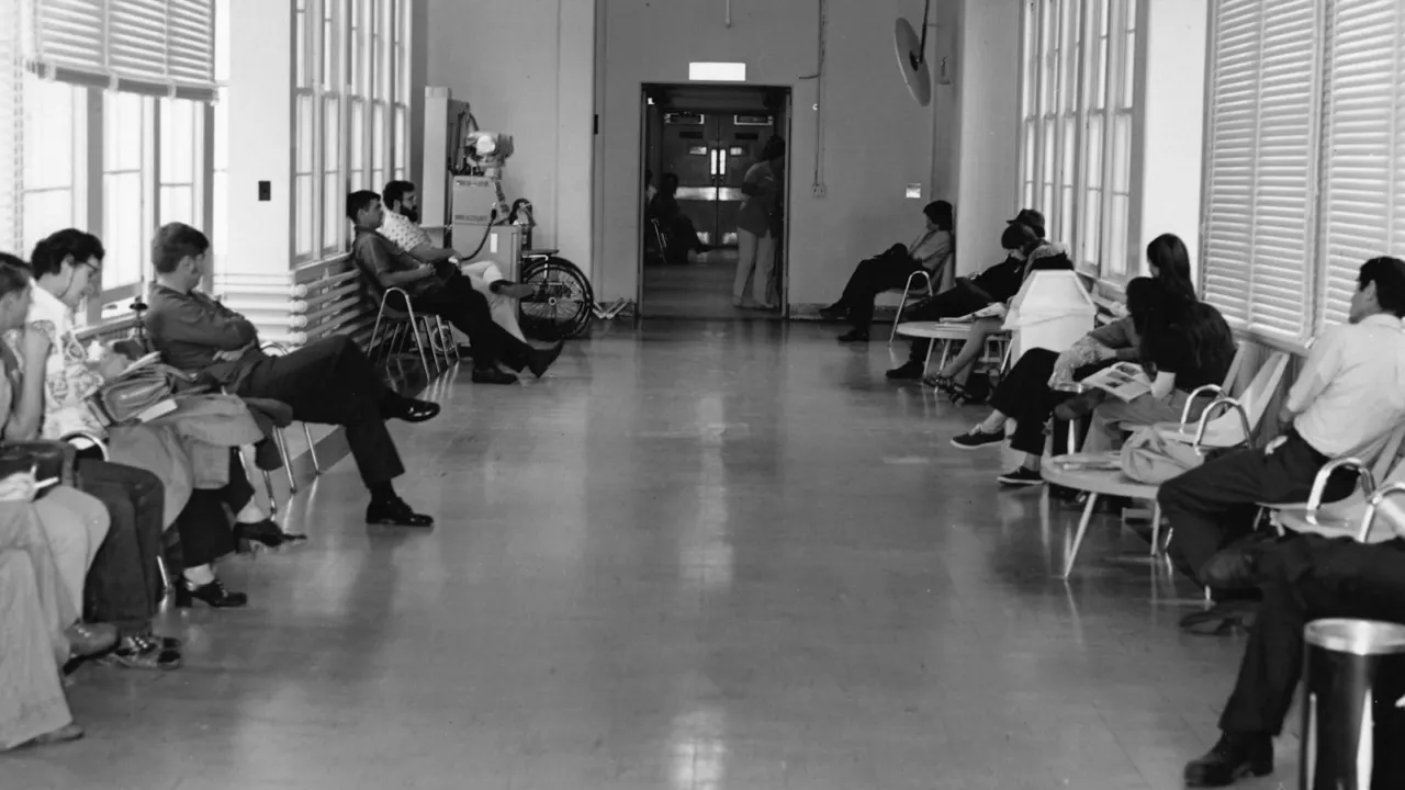 Black and white photo of patients seated on chairs along both sides of a long clinic corridor