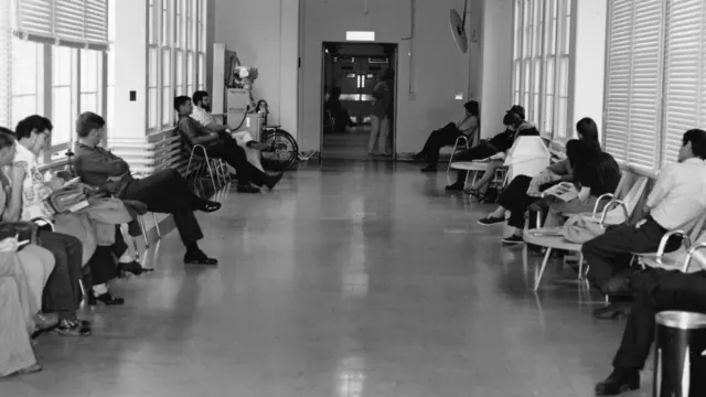 Black and white photo of patients seated on chairs along both sides of a long clinic corridor