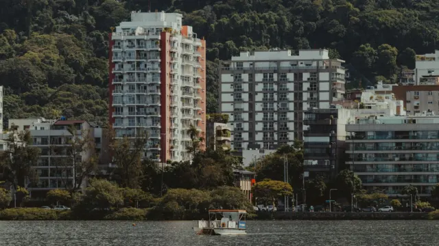Apartment towers lining a calm waterfront with a small boat on the water and green hills behind