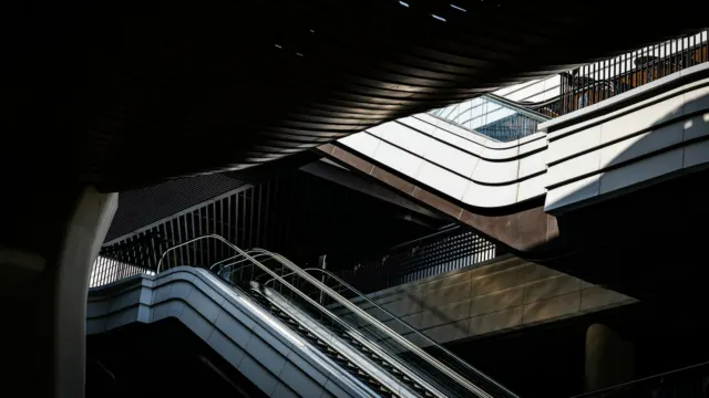Escalator rising through a bright mall interior under a skylight casting light and shadow